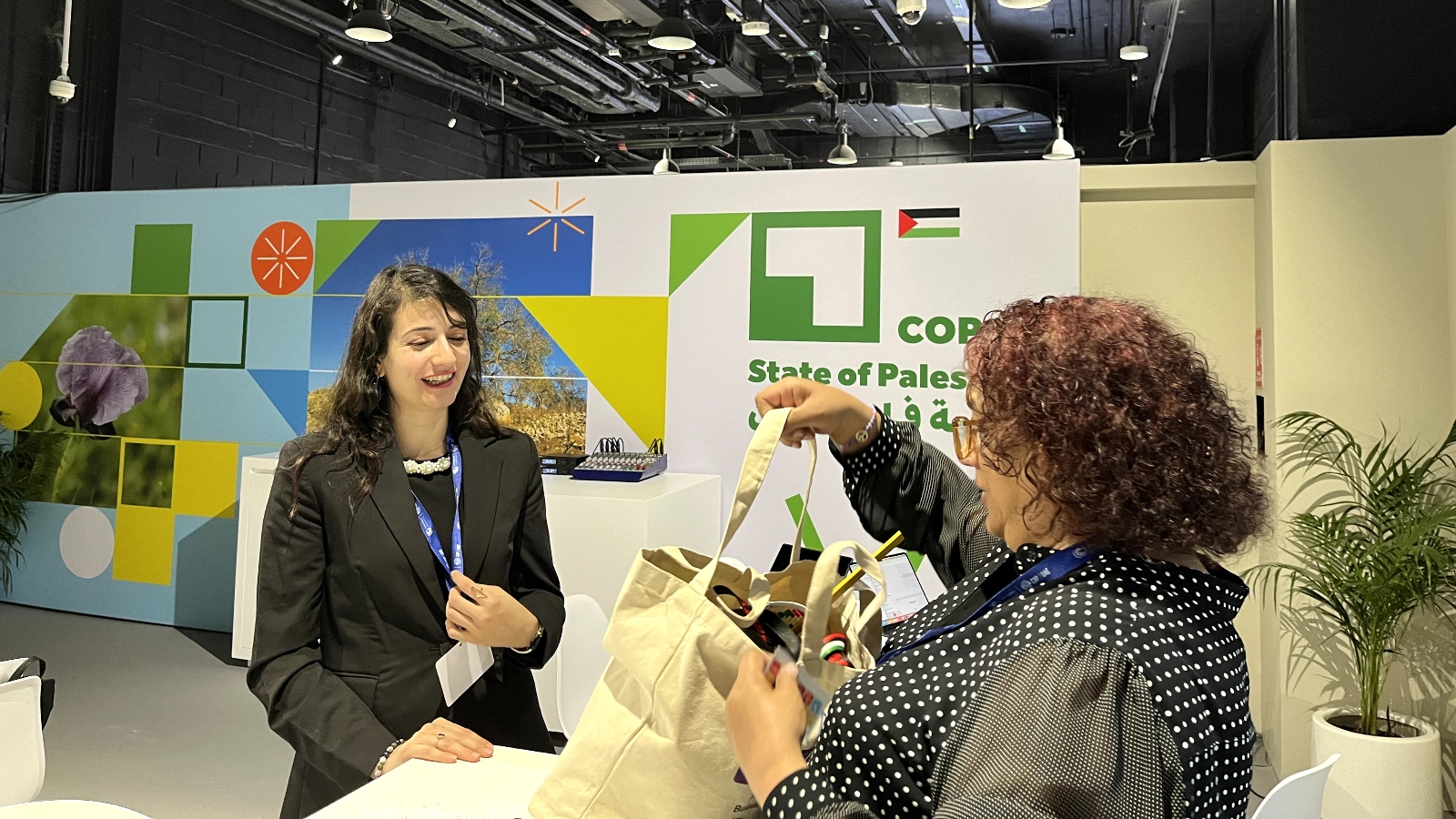 A smiling woman in a black suit faces another woman holding a tote bag in front of a sign that says "COP ... State of Palestine"