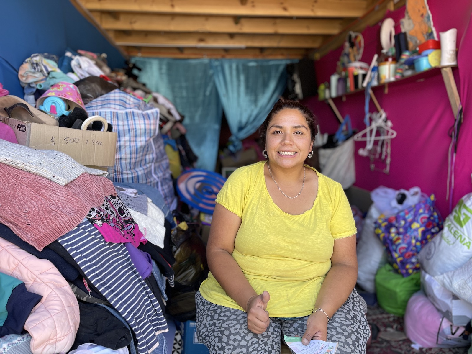 a smiling woman in a yellow shirt sits in a small room with a large pile of clothing