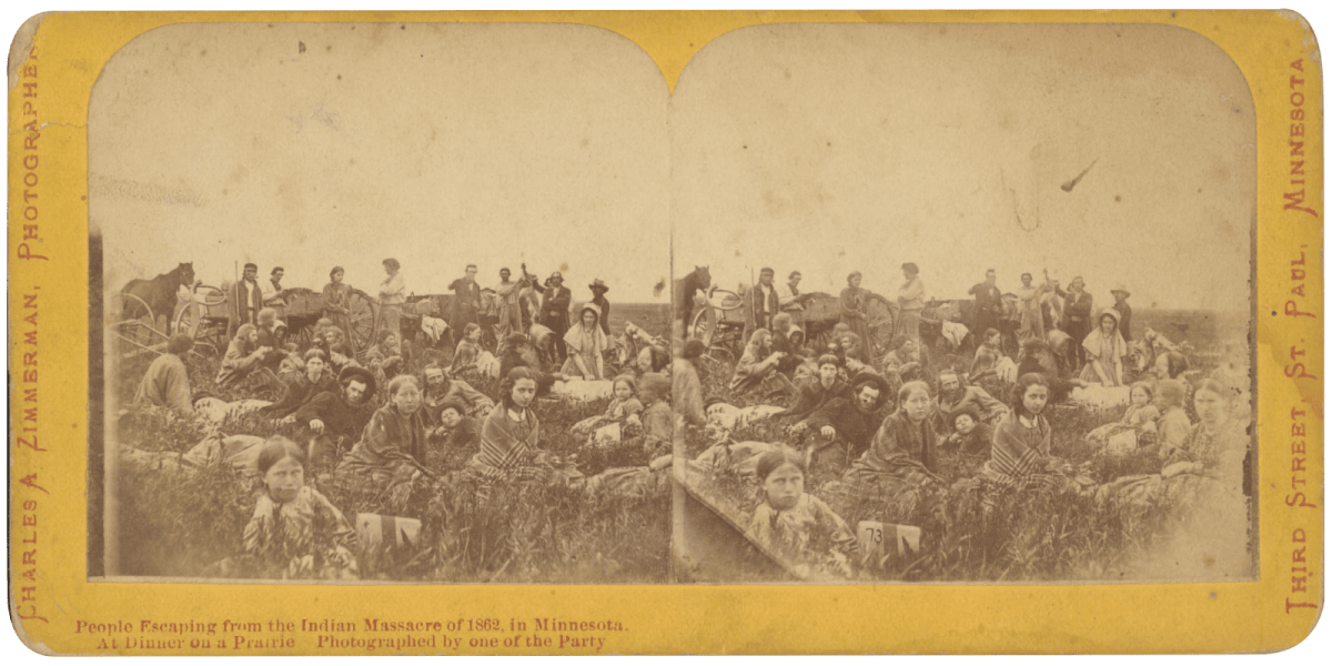 Outdoor group portrait of refugees of the Dakota War of 1862 gathered on prairie for meal.