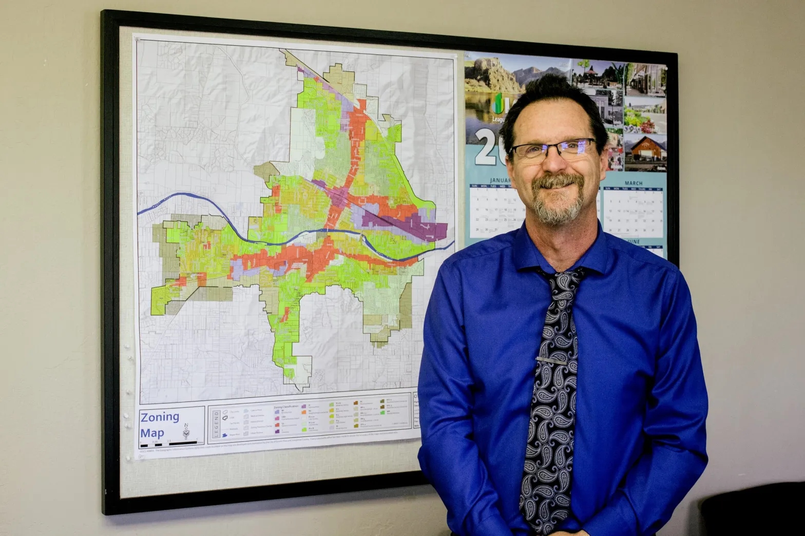 A man with a goatee and glasses, wearing a bright blue button down shirt and black tie, stands in front of a map.