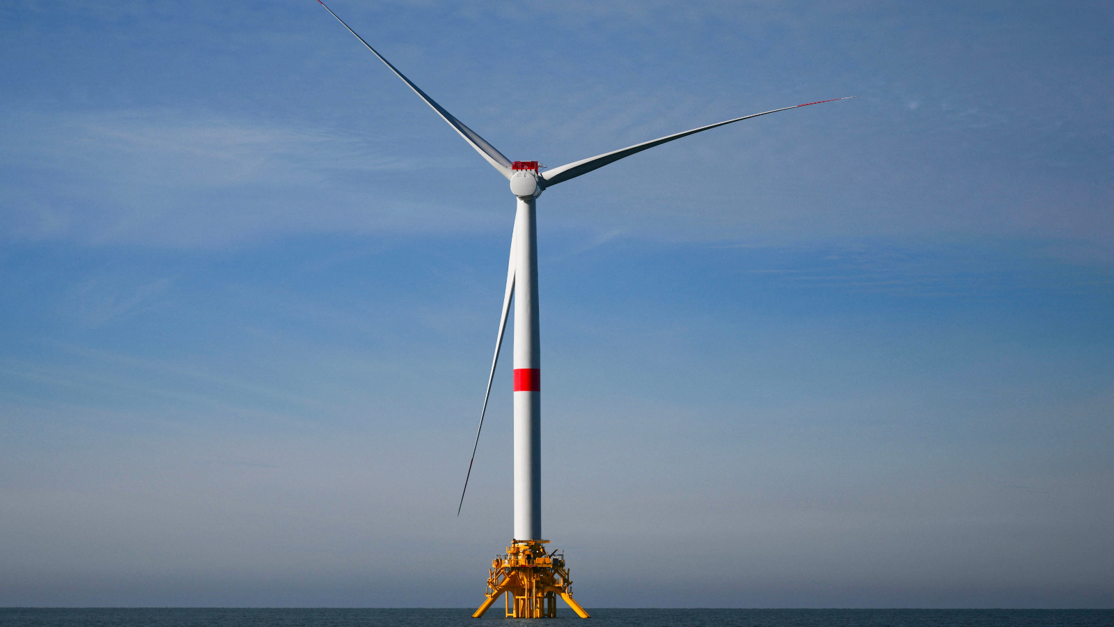 An offshore wind turbine, mostly white with a red stripe around the middle of its tower, against a partly cloudy sky