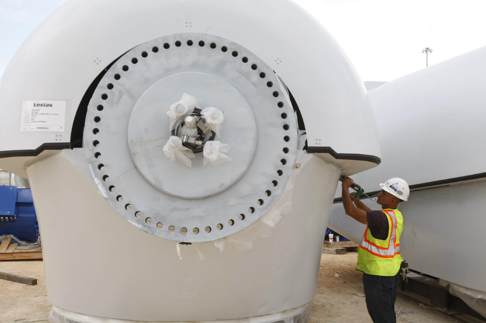 Man wearing bright yellow vest and white helmet holds a tool up to a large white piece of machinery about twice his height