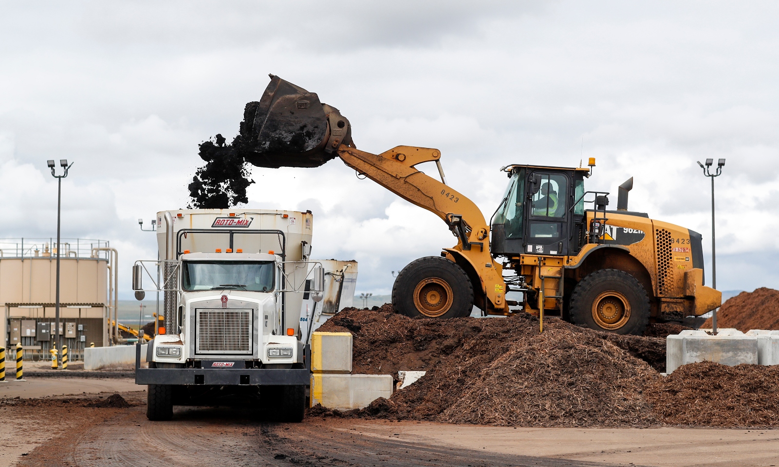 A yellow excavator dumps brown, earthy material into the back of a white mixing truck