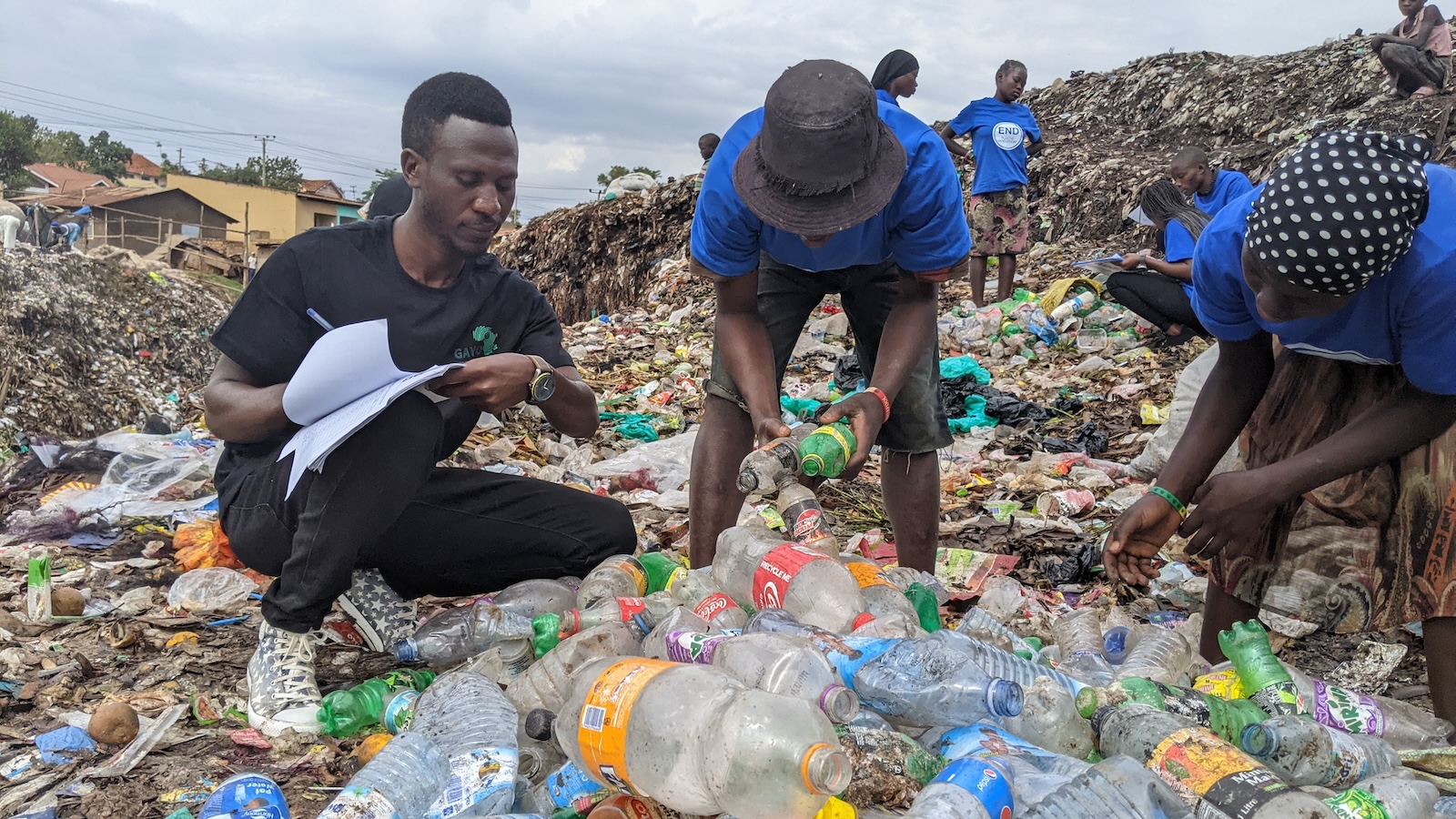 Three people bend over pile of collected plastic items, with trash in background