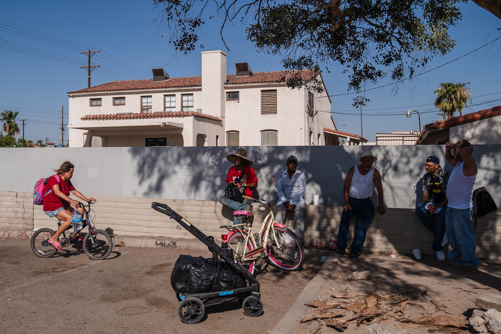 a group of men in the shade of a tree along a sidewalk. A person rides a bike toward them while carrying a plastic gallon water bottle