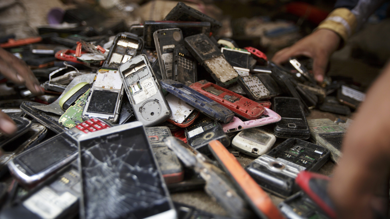 A hand reaches into a large pile of cell phones and other small electronic devices, many of them cracked or missing batteries