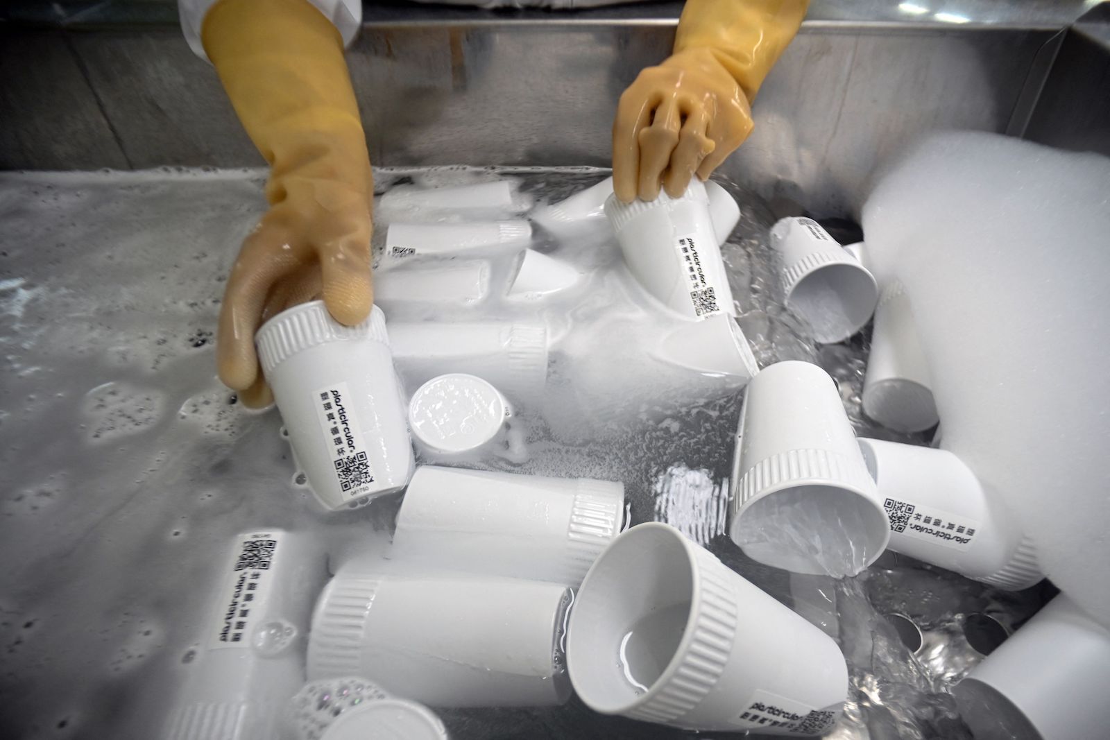 Hand with yellow rubber gloves wash many white plastic cups in a large basin of water