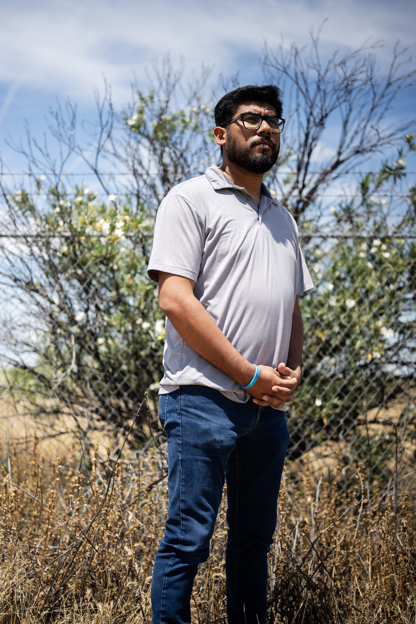 A man stands with hands clasped near a bush