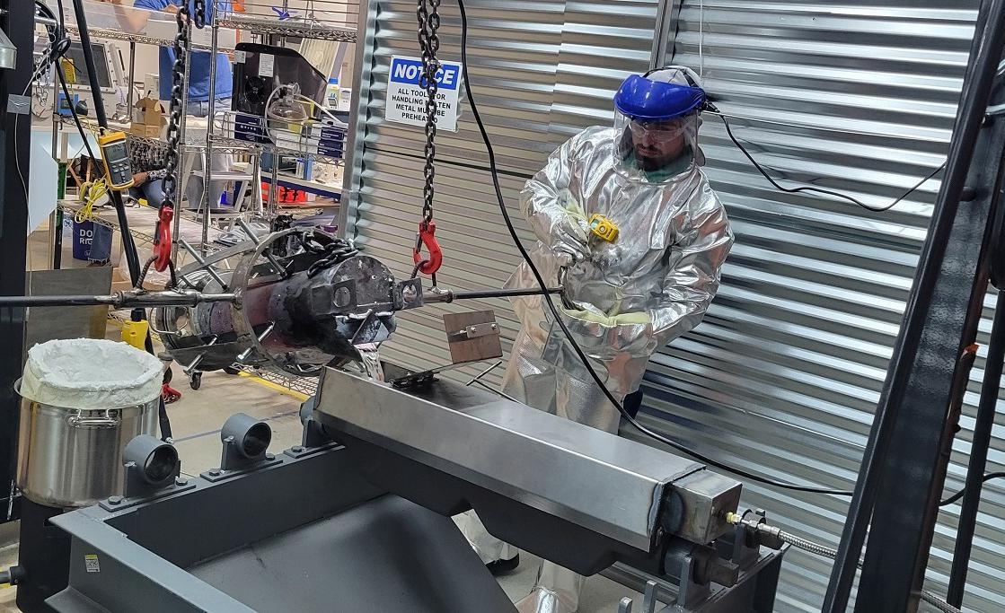 A man in a full length silver suit, helmet, and face shield works with heavy-duty equipment.