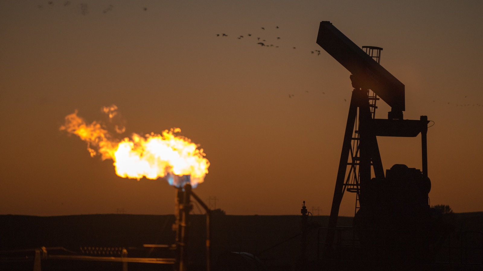 A pumpjack in the foreground and a burning flare in the background of an oil field in Montana, with dark orange sky.