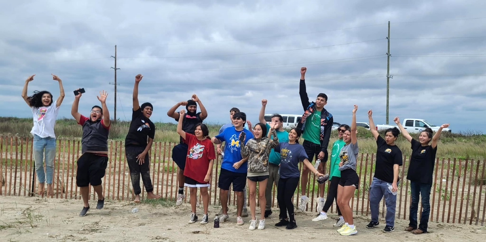 A group of teenagers stand near a fence. They are cheering and jumping.