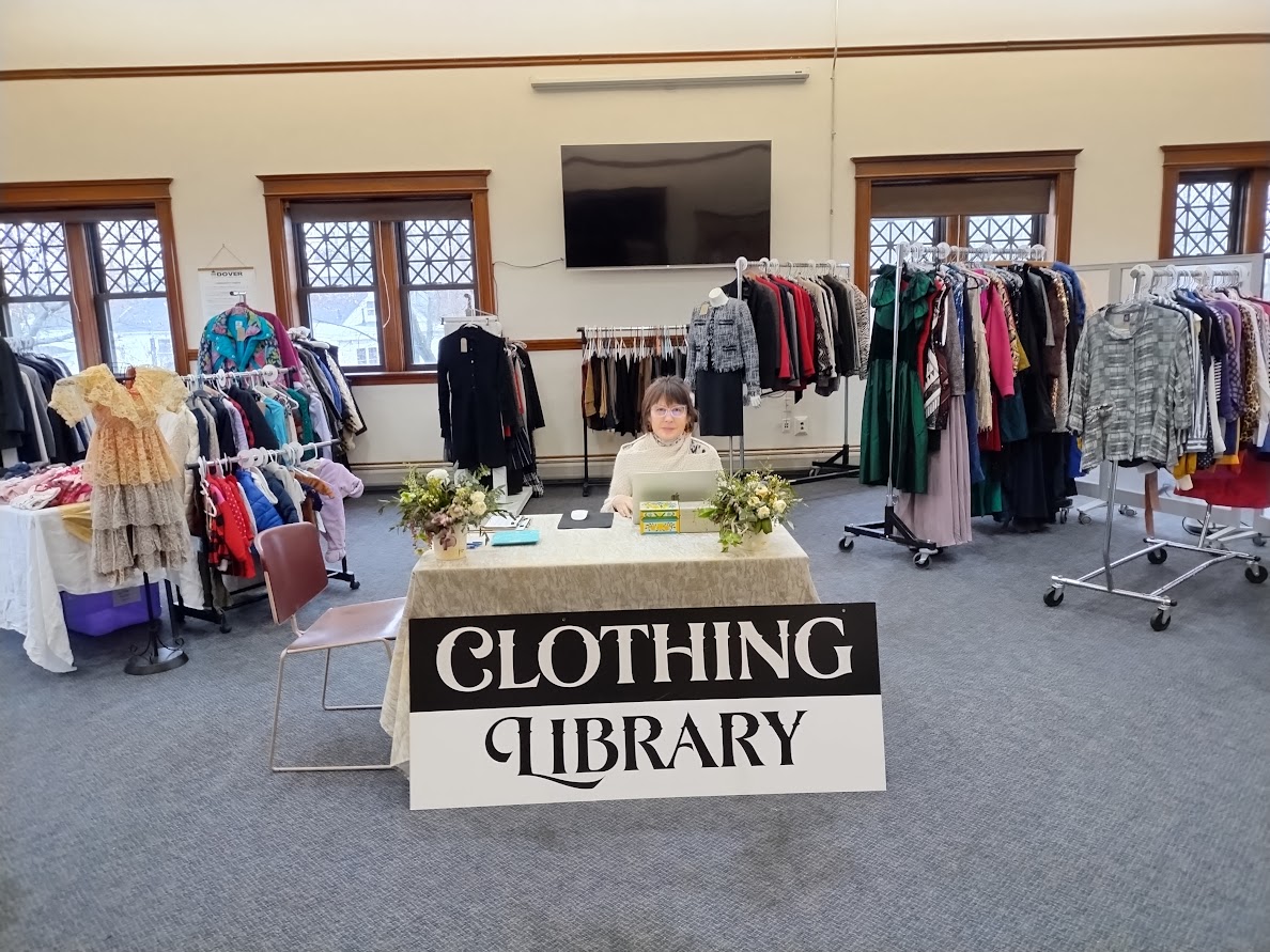 A woman sits at a table in a large meeting room, surrounded by racks of clothes on hangers