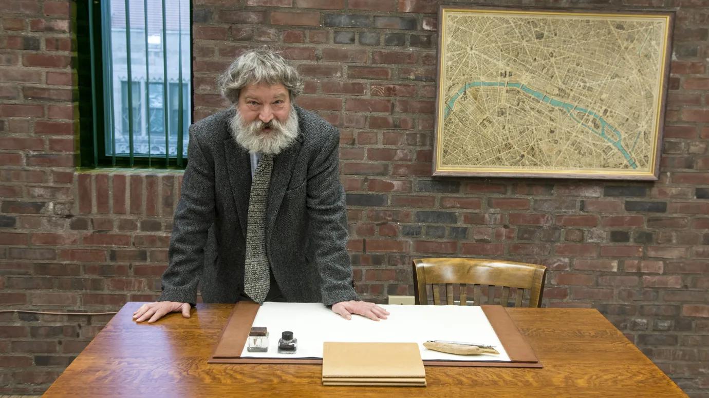 A man with a mop of gray hair and a long gray beard stands in a gray suit jacket with his arms on a desk.