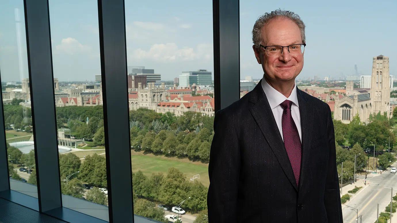 A man in a suit and glasses stands in front of floor to ceiling windows.