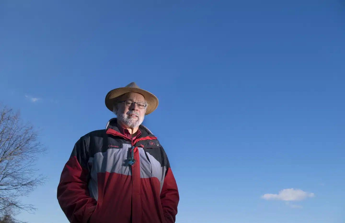 A man in a red jacket and straw hat stands in front of a blue sky.