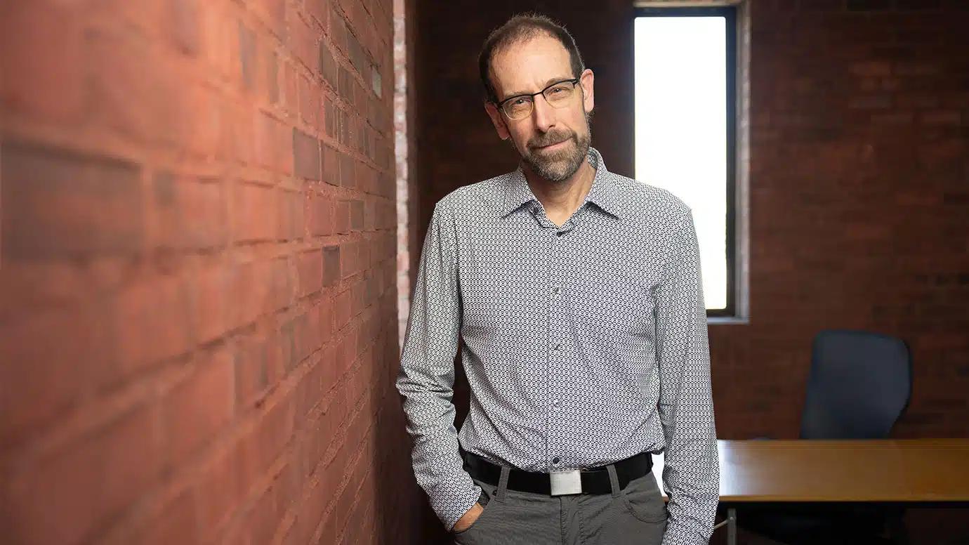 A skinny man with a goatee and glasses wears a gray button down shirt and stands near a wall.