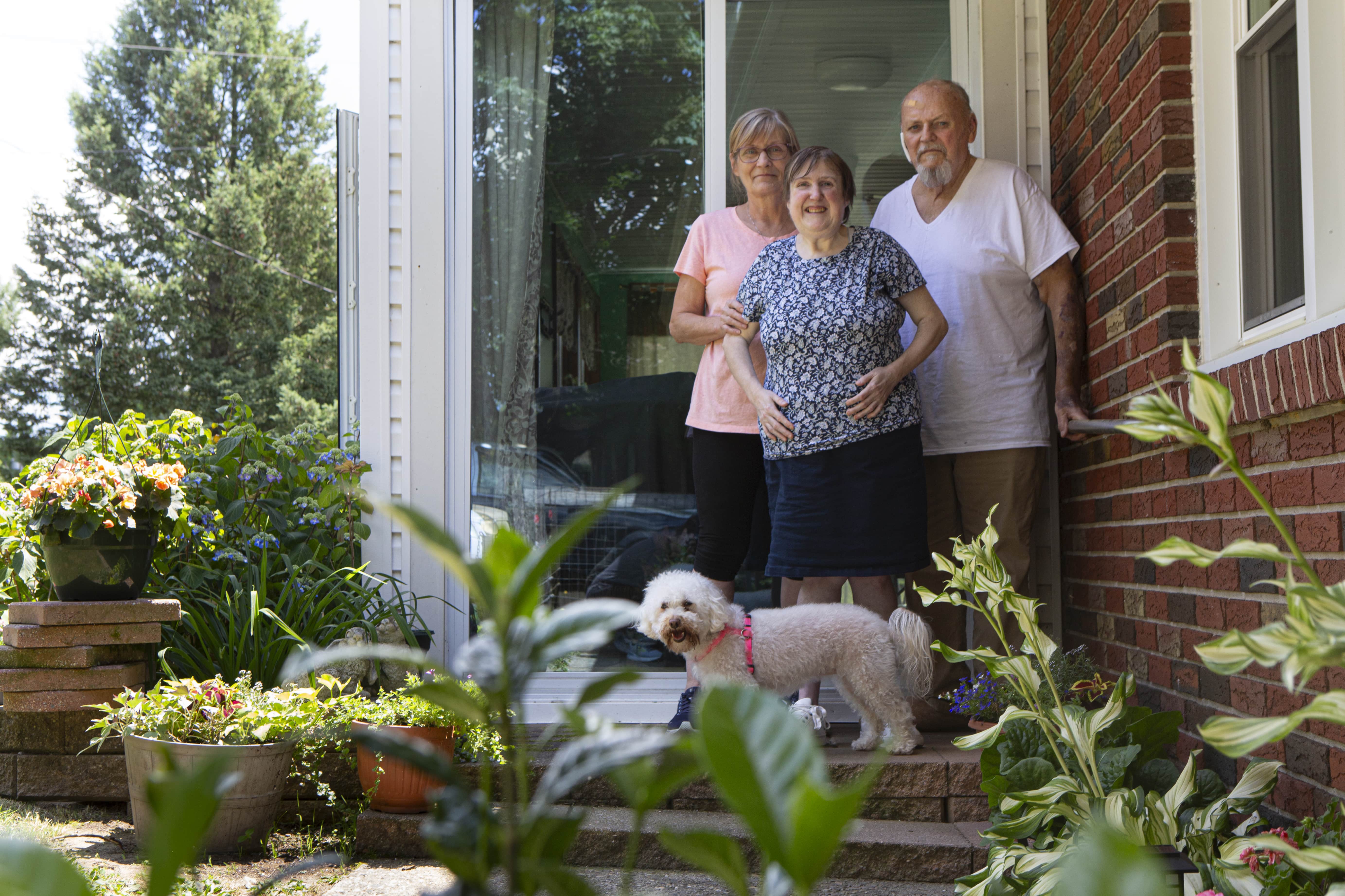 Two women and a man stand with a fluffy white dog outside of a house with lots of plants.