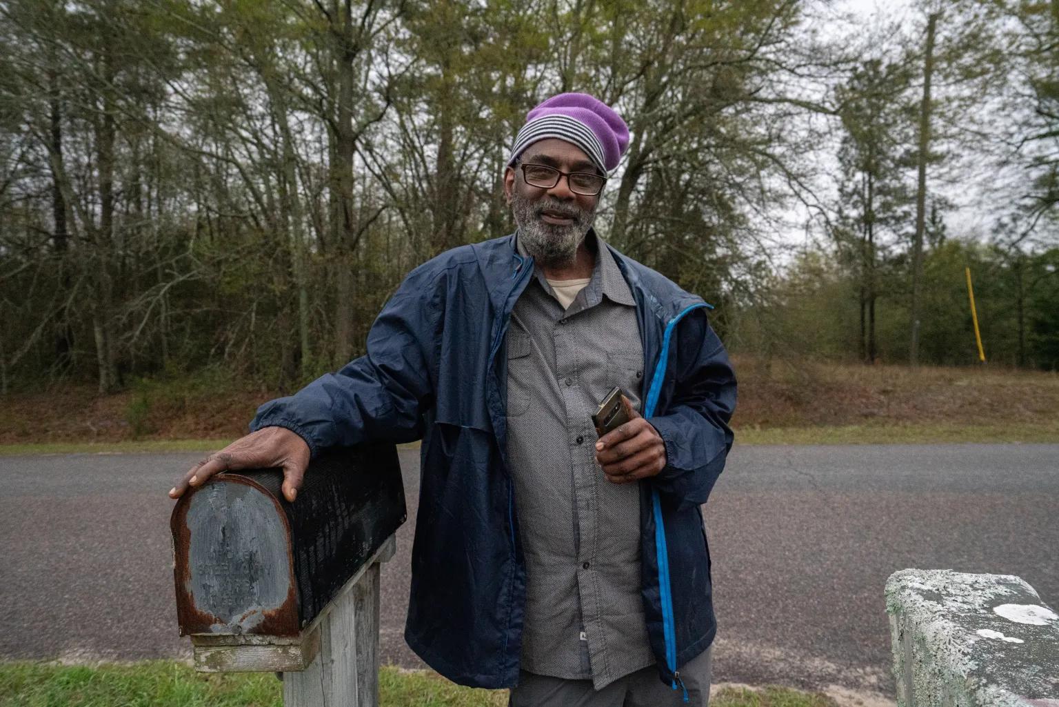 A man wearing glasses in a gray button-down shirt, blue jacket, and purple hat smiles while leaning on a mailbox.