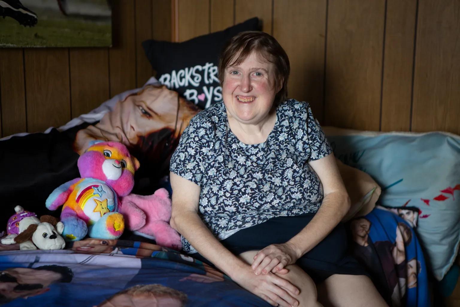 A woman in a blue flowered shirt and skinny arms sits on a bed, smiling.