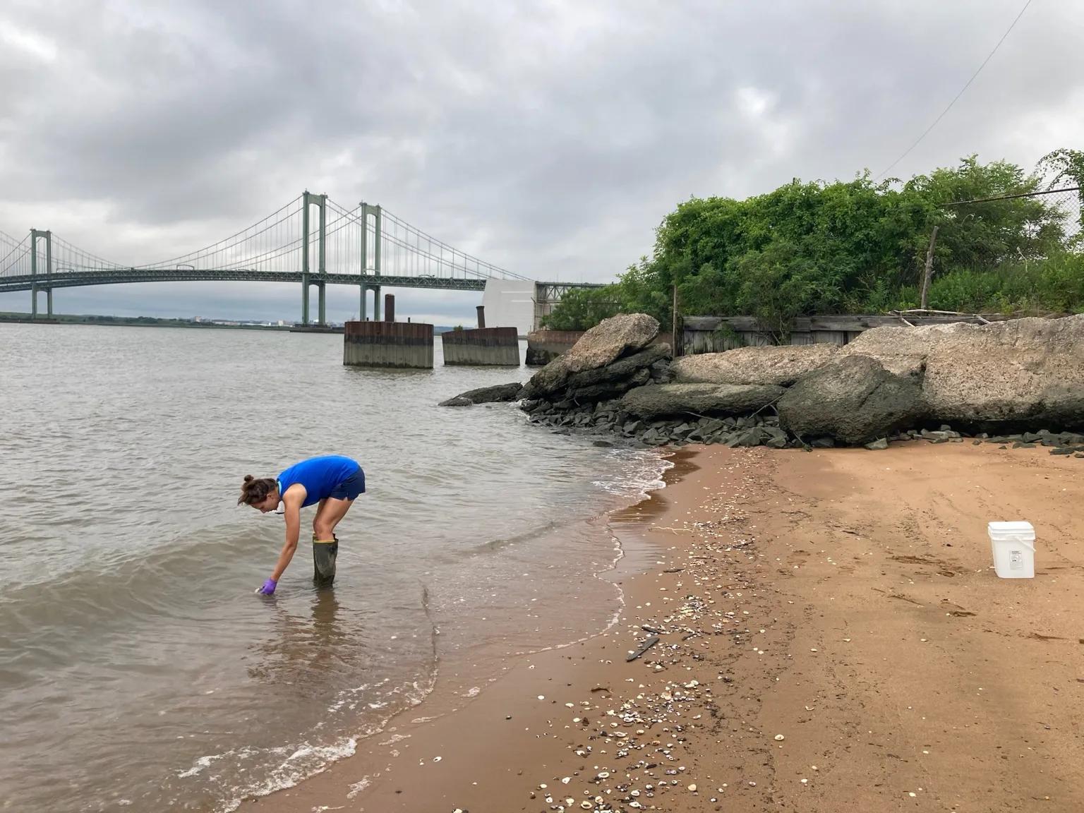 A woman in rain boots and a blue shirt bends down to collect river water on the edge of the water.