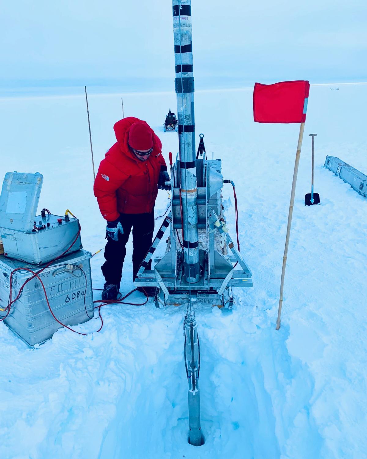 A person in a bright coat and hat stands next to a tall cylindrical device drilling into a hole in the ice