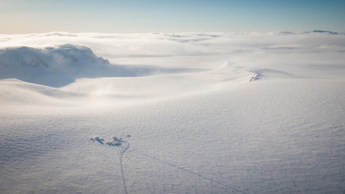 An aerial photo of a huge expanse of snow and ice, with a tiny cluster of tents visible in the bottom left corner, and snowmobile tracks extending out from it