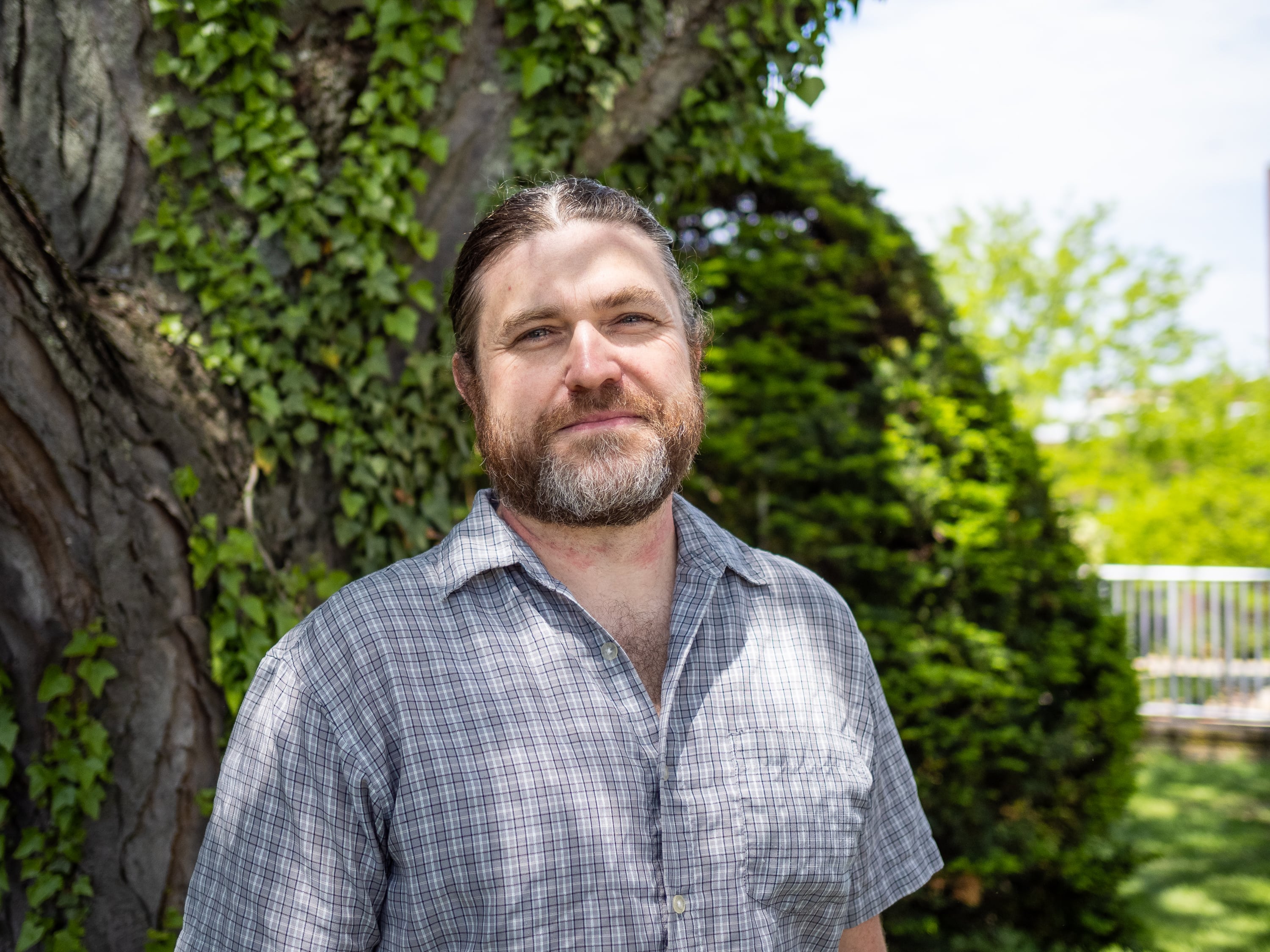 A bearded man with long hair tied back stands in front of a tree.