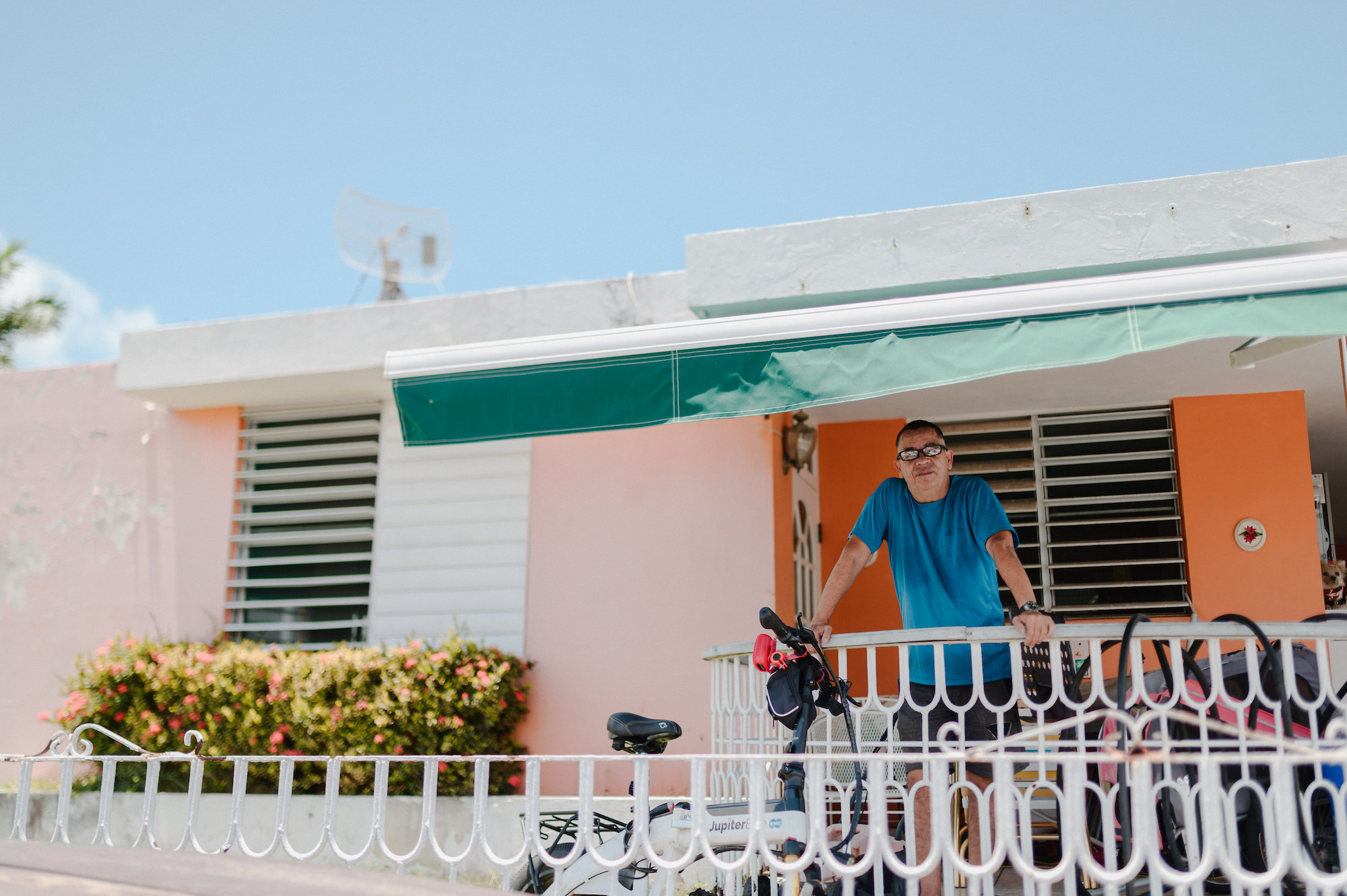 A man in a teal shirt stands on the deck of a colorful pink house