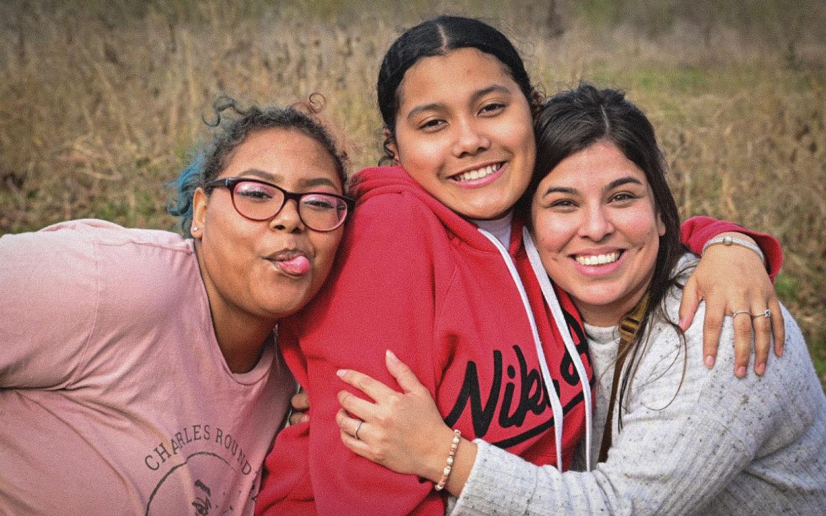 A woman and two teenage girls embrace, smiling at the camera