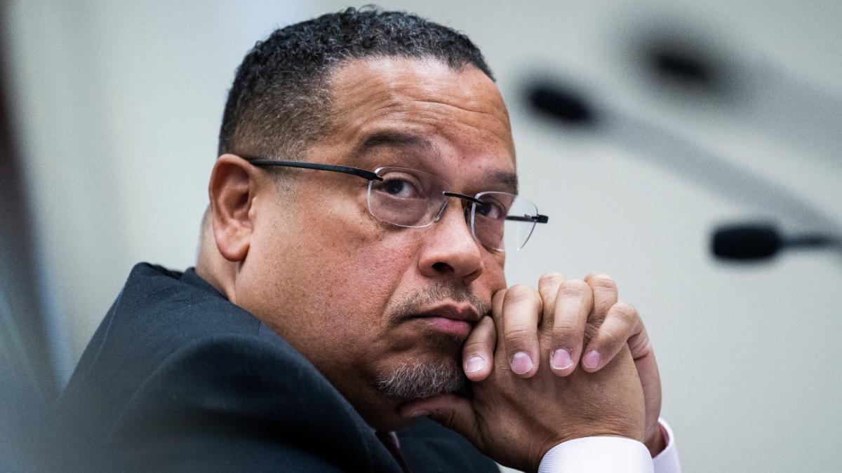 A man with short black hair, a beard, and glasses stares into the distance.