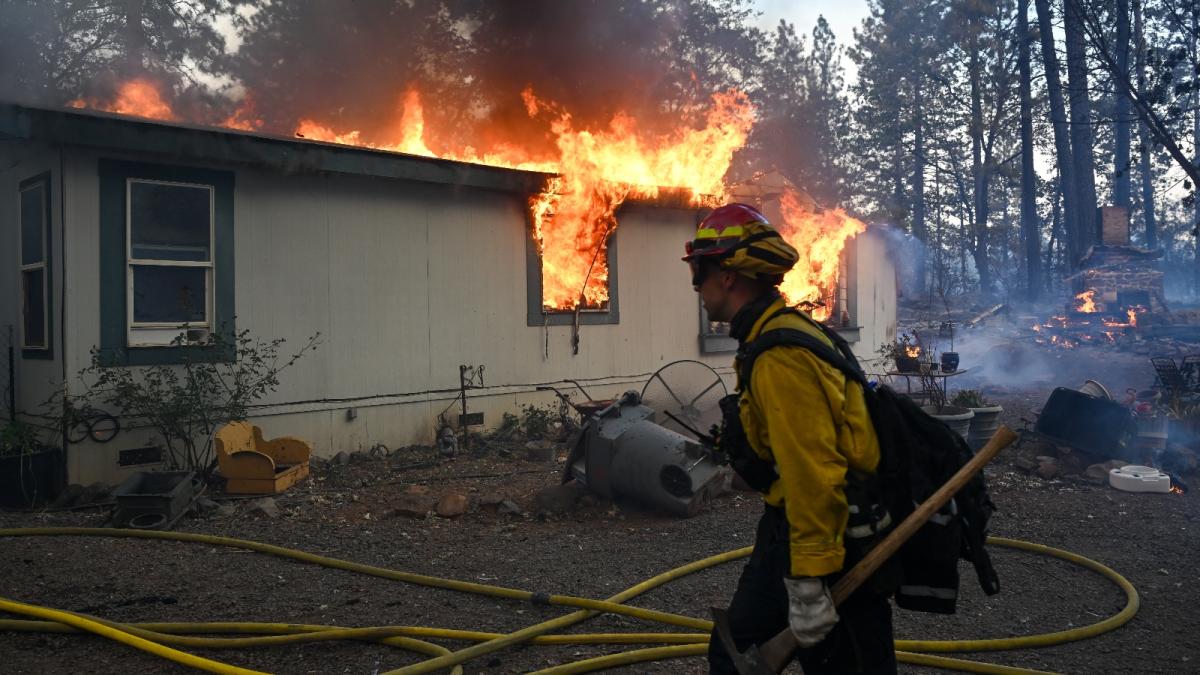 A firefighter with an axe in his hand walks in front a house on fire.