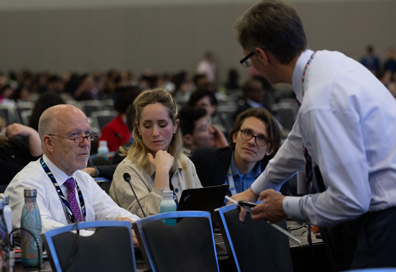 Three people seated at a table interact with a man on the other side of the table. Behind them are many others, out of focus.