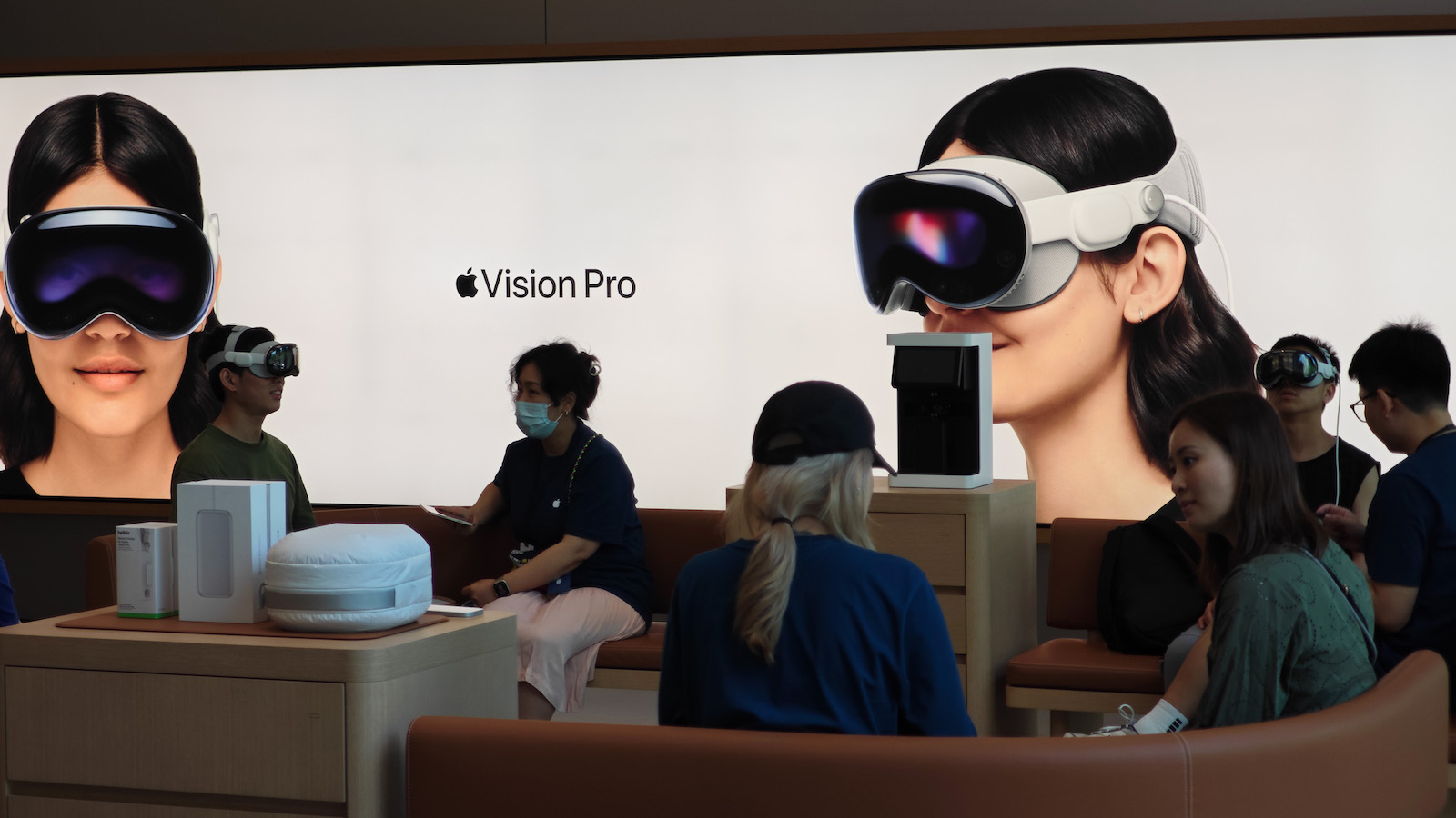 Customers mill about in front of a large white screen with a photograph of a smiling woman wearing a VR headset and the text 'Vision Pro' next to the Apple logo