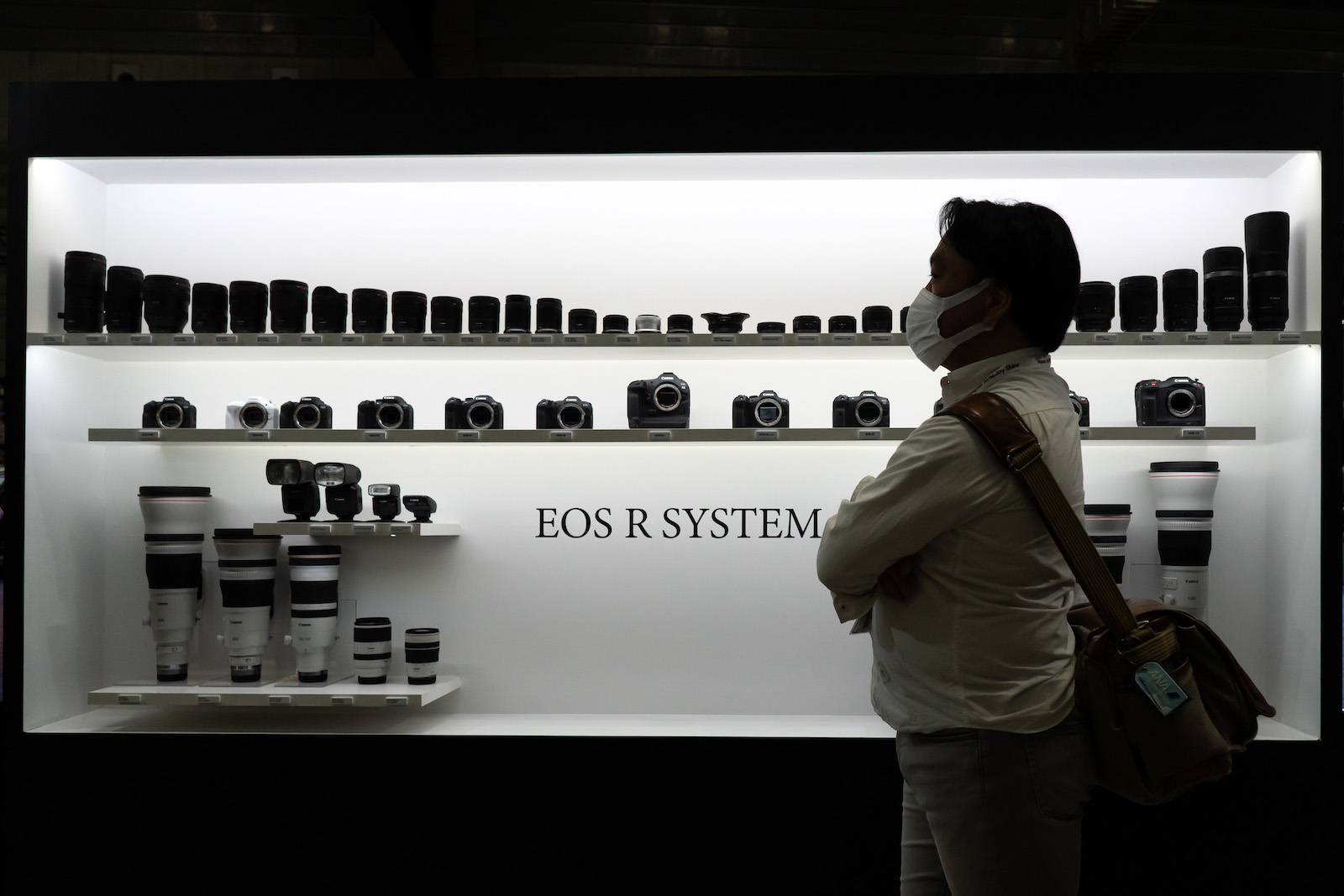 A person wearing a masks stands in profile in front of a sleek display of small cameras and equipment, which appear black against a white background that bears the text 'Eos R System'