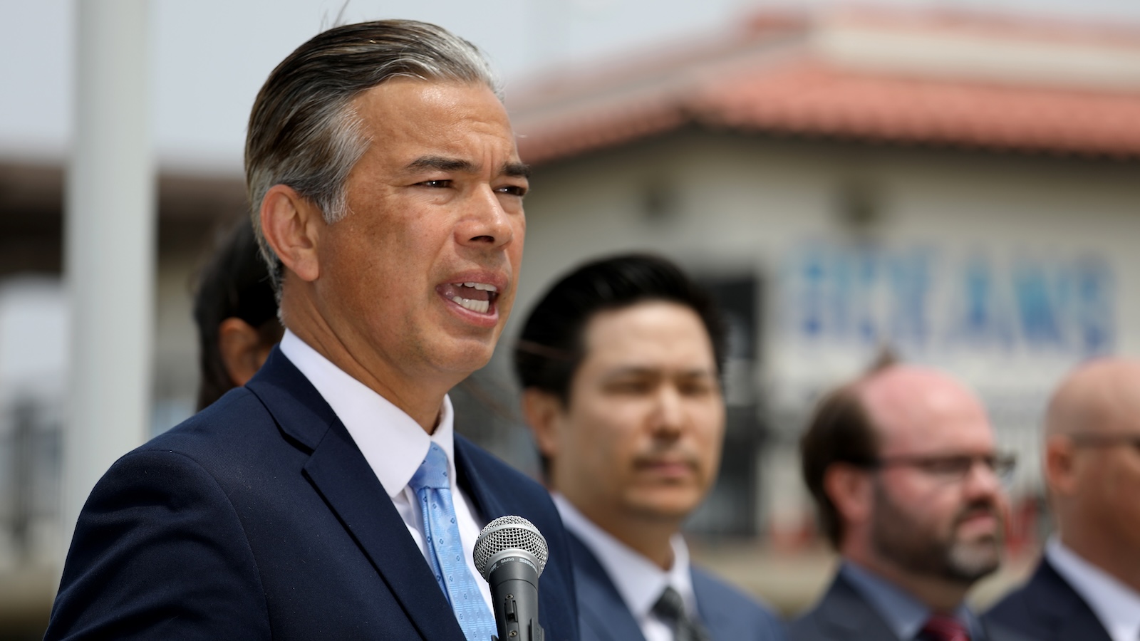 Side view of California Attorney General Rob Bonta speaking at a podium, flanked by several men in suits.