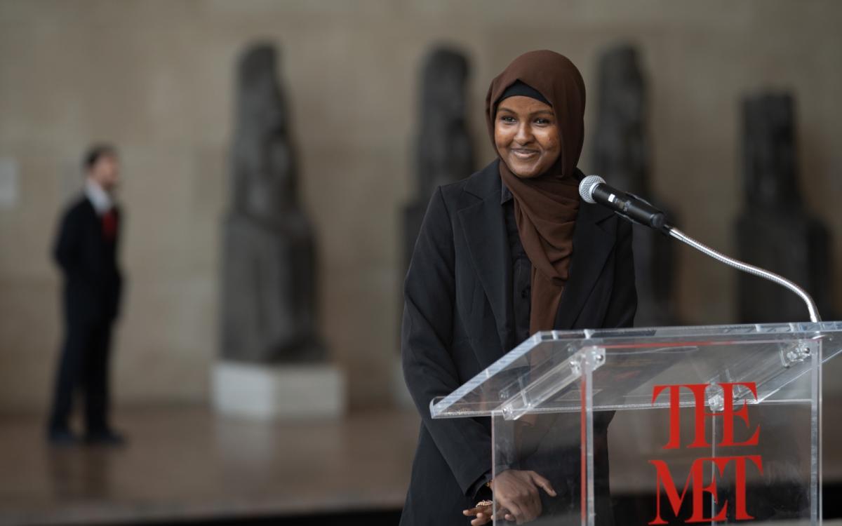 A young woman wearing a head scarf smiles, standing in front of a podium labeled "THE MET" with a microphone