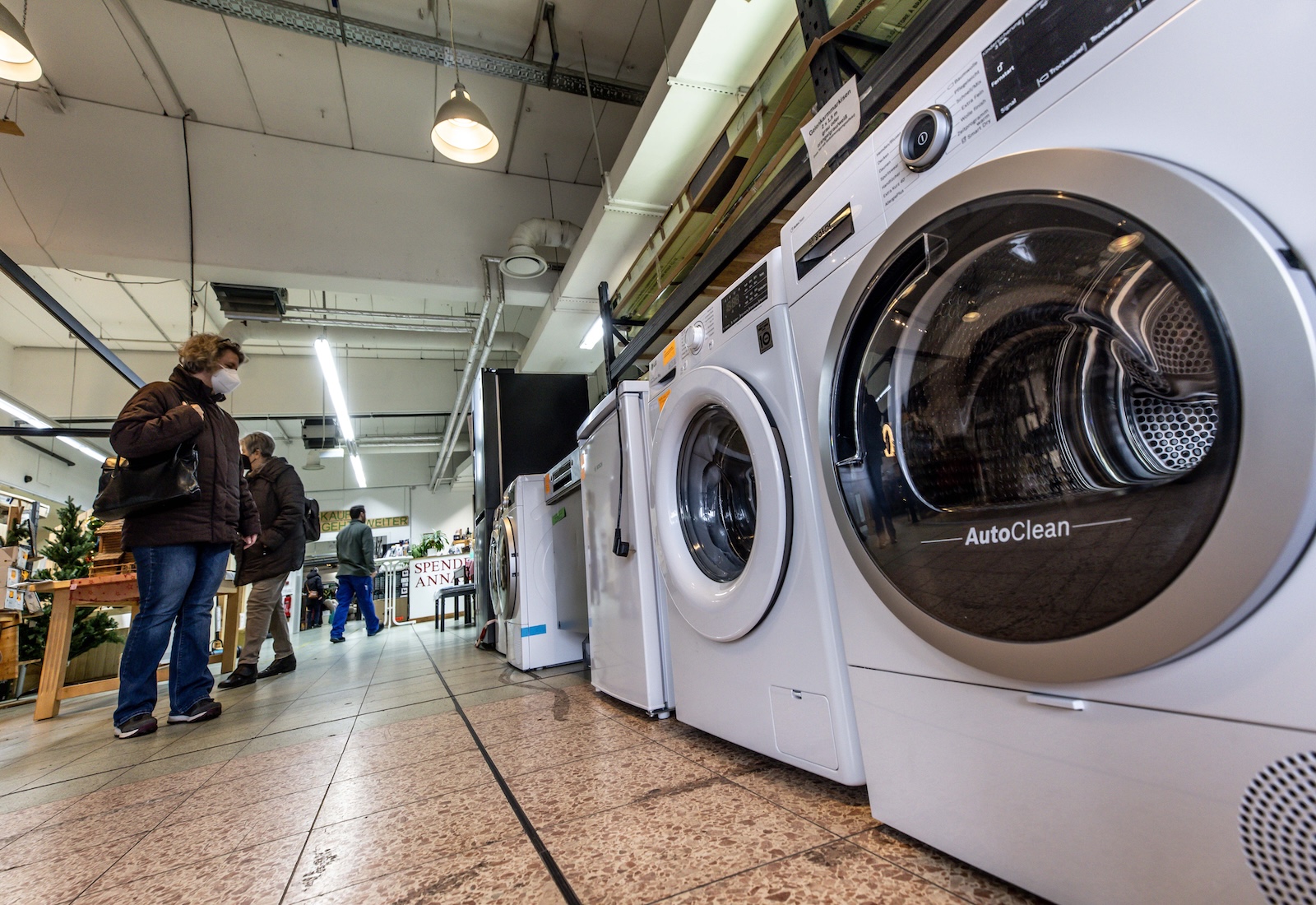 White washing machines lined up on a row on a shopping room floor, with shoppers looking at them.