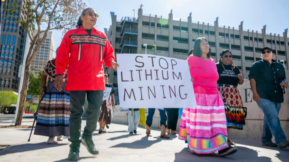 A man in a red sweater and glasses carries a Stop Lithium Mining sign which is also held by a woman in a pink shirt and skirt