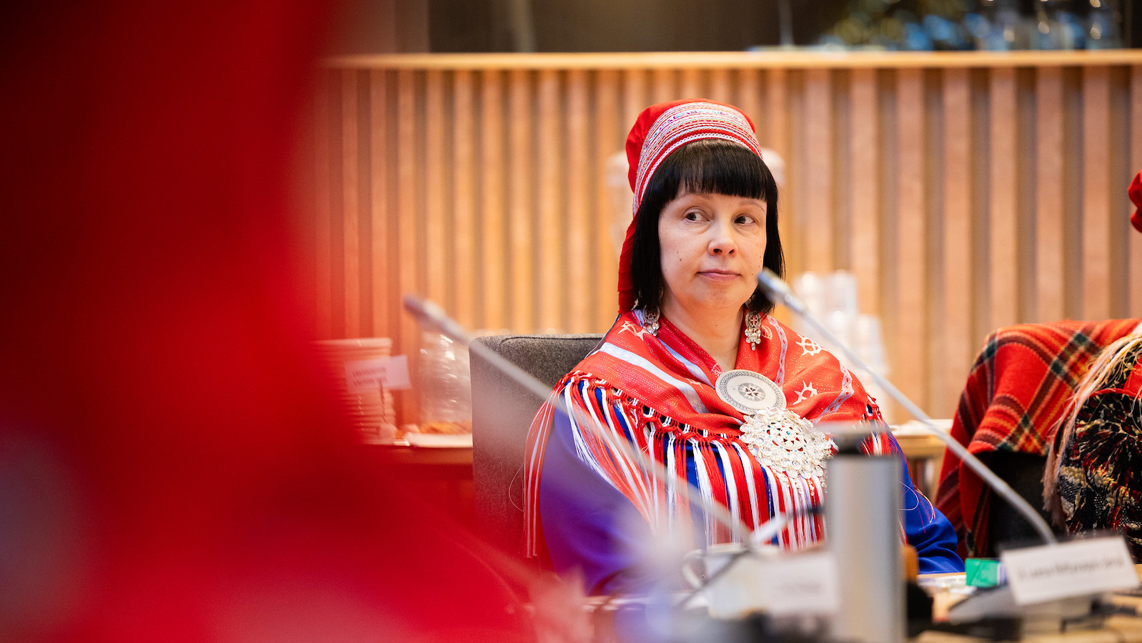 A woman in traditional Sámi dress -- a red, white, and blue shawl tied at the neck with an elaborate silver clasp and matching head covering -- sits at a conference table with a microphone. She looks off to the side as if listening to someone.